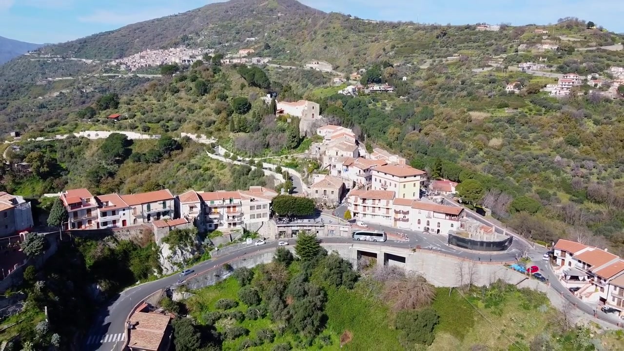 Ripresa aerea con allontanamento che rivela il borgo medievale collinare di Savoca in Sicilia, circondato da lussureggianti montagne mediterranee, Bar Vitelli in Piazza Fossia, famoso per essere stato un luogo di riprese de Il Padrino.