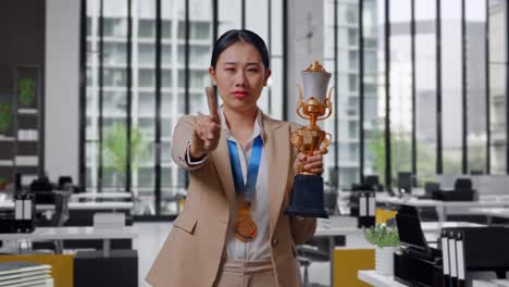 asian business woman in a suit with a gold medal and trophy disapproving with no index finger sign and smiling to camera in the office