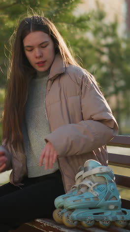 una joven con una chaqueta de melocotón, camisa interior gris y pantalones negros se sienta en un banco del parque. coloca sus patines a su lado y descansa sus manos en su barbilla