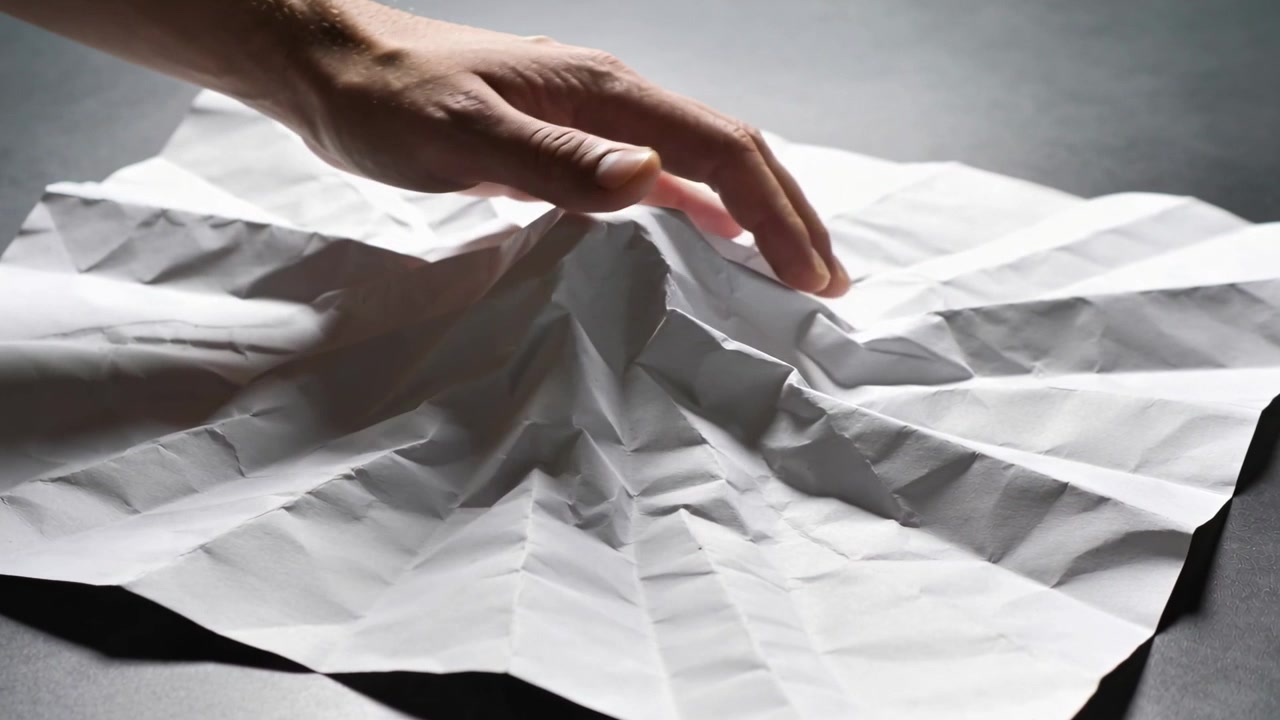 Touching center, adult hand pinching, lifting crumpled paper peak at dark tabletop with soft light