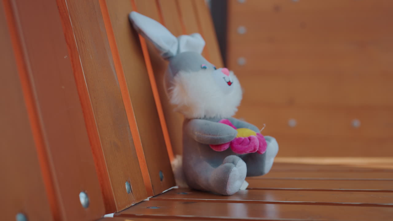 Panchina del parco: coniglietto di peluche rubato. Una mano guantata afferra rapidamente il giocattolo e lo porta via. Movimento affrettato catturato in primo piano, posto vuoto lasciato indietro, tesa mattina urbana, azione di scippatore/ladro di strada.