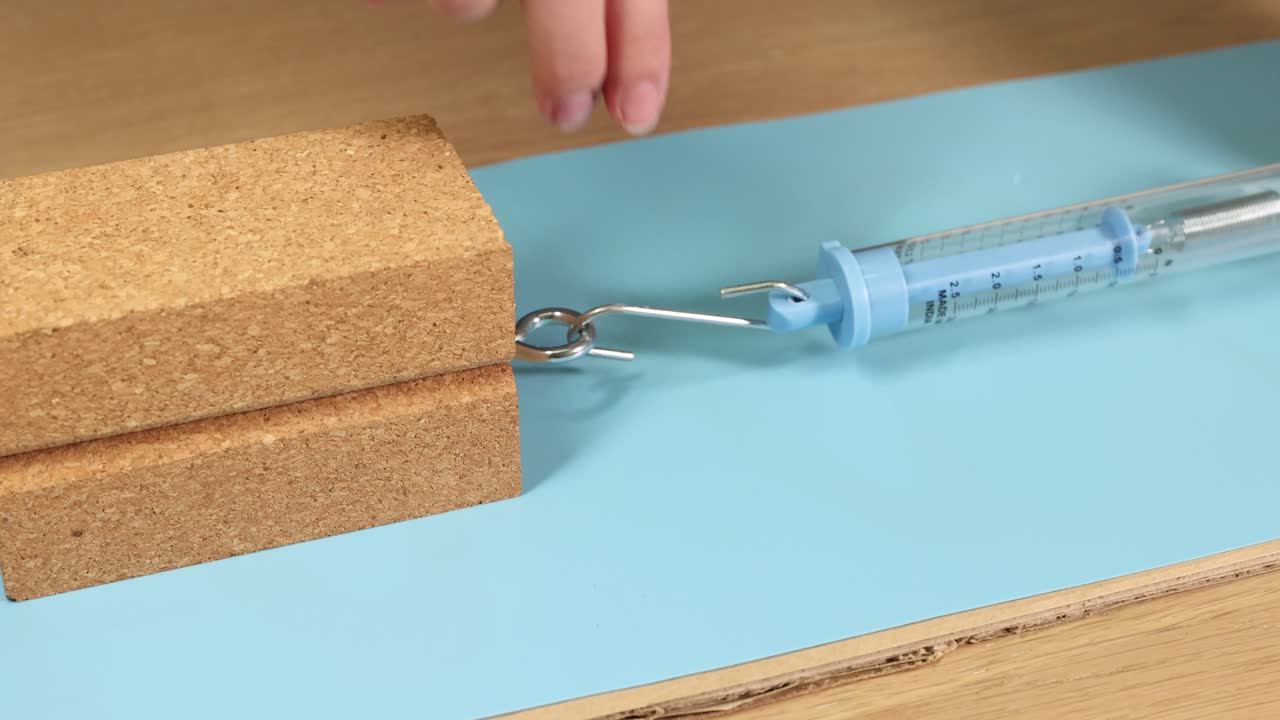 A hand uses a spring scale to measure friction on a wooden block. Bright lighting and close-up focus enhance the experiment