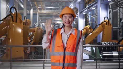 asian female engineer with safety helmet smiling to camera and waving hand saying bye while standing with industrial robotic arms working
