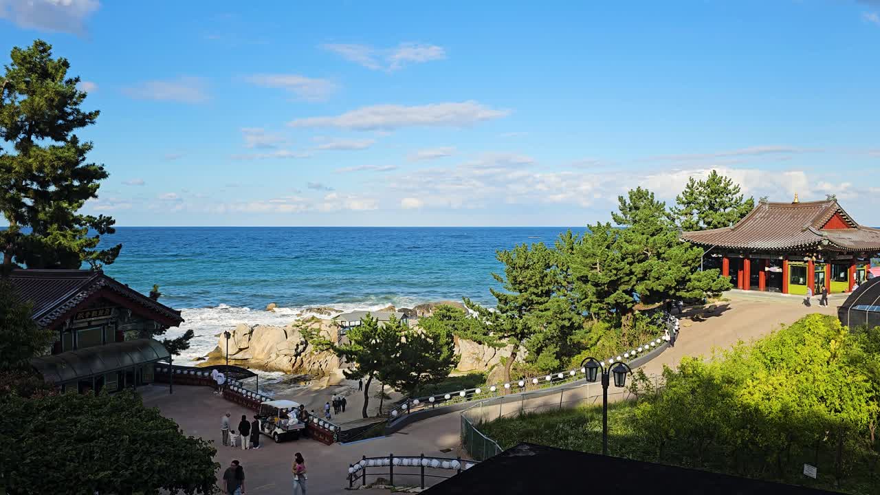 Wide angle overview of Gangwon Hyuhyuam Hermitage along the coastline, showcasing the peaceful ocean and the temple nestled in the cliffs, tourists admiring ancient Korean architecture in summer