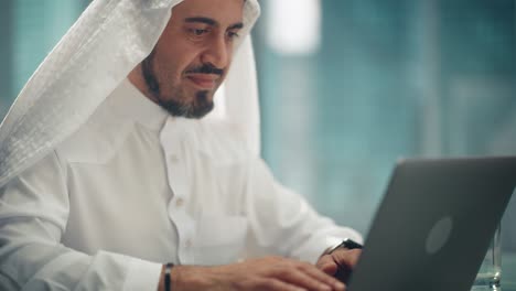 empresario árabe exitoso con traje tradicional blanco sentado en la oficina y trabajando en una computadora portátil. gerente de negocios planeando estrategia corporativa. saudí, emiratí, concepto de empresario árabe.