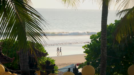 Blick-Von-Der-Bali-Lounge-Durch-Kokospalmenwedel-Am-Strand-Von-Seminyak-Bei-Sonnenuntergang-Mit-Nicht-Erkennbaren-Menschen,-Die-Am-Sandstrand-Spazieren---Zeitlupe