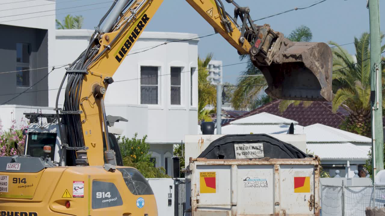 Excavator transfers large dirt load into dump truck, bright daylight, suburban residential construction environment