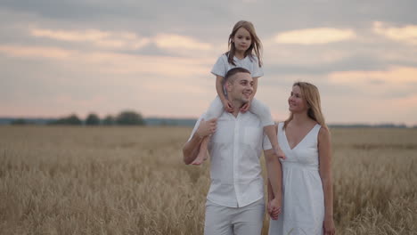 Young-couple-of-parents-with-girl-children-holding-hands-of-each-other-and-running-through-wheat-field-at-sunset.-Happy-family-jogging-among-barley-meadow-and-enjoying-nature-together.-Slow-motion