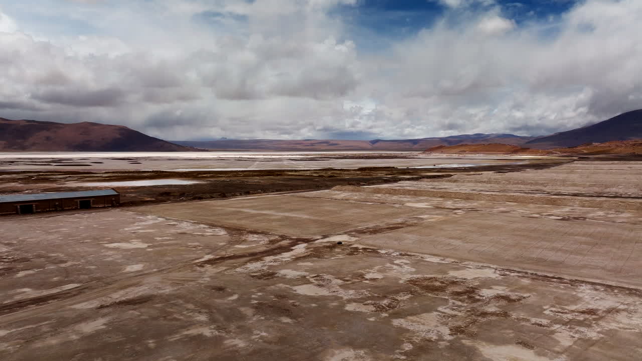Drone view of remote Bolivian borax mine on arid plains near Laguna Capina, capturing the barren salt-crusted landscape, isolated structure, and distant mountains under cloudy skies