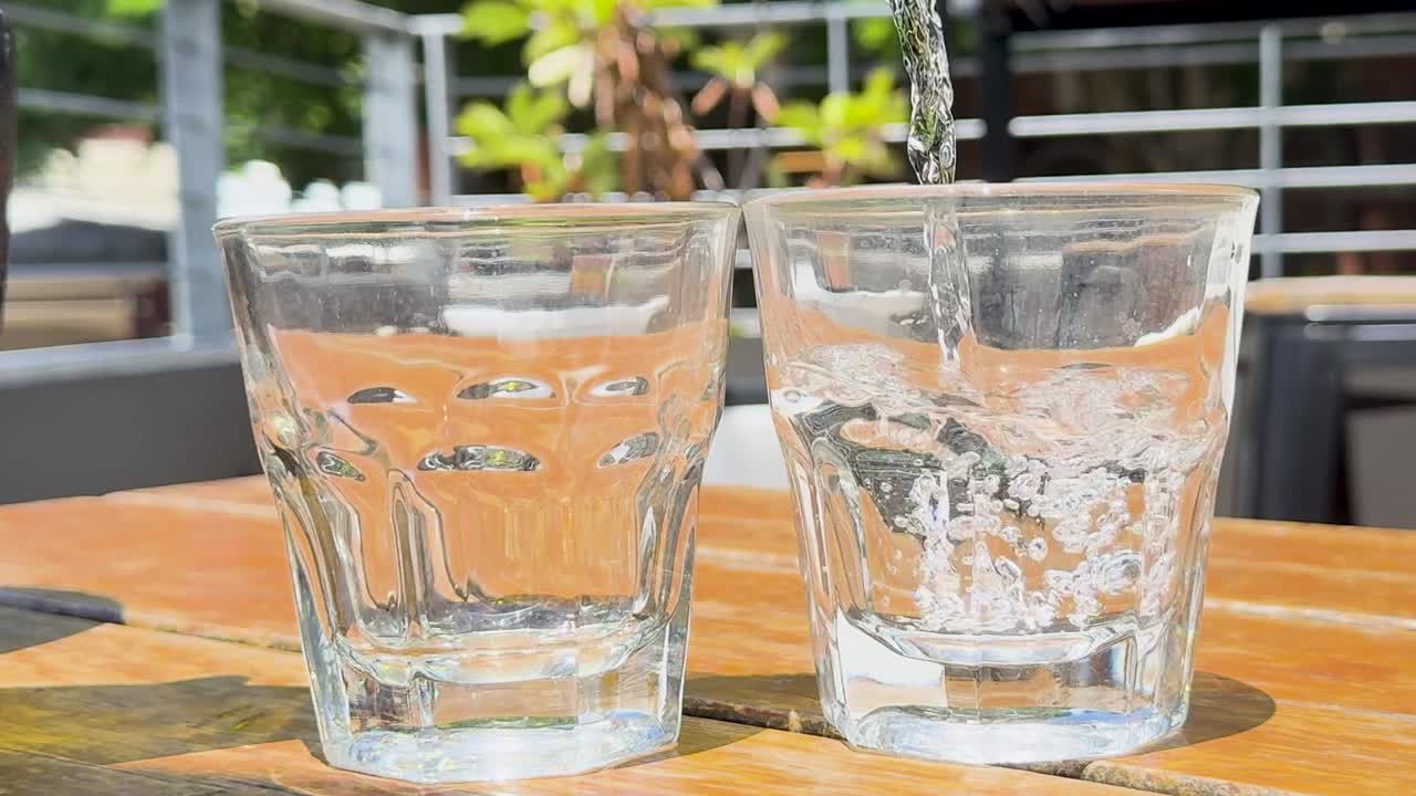 Nước mát làm đầy hai cốc thủy tinh có họa tiết đặt trên một chiếc bàn gỗ ngoài trời được chiếu nắng.