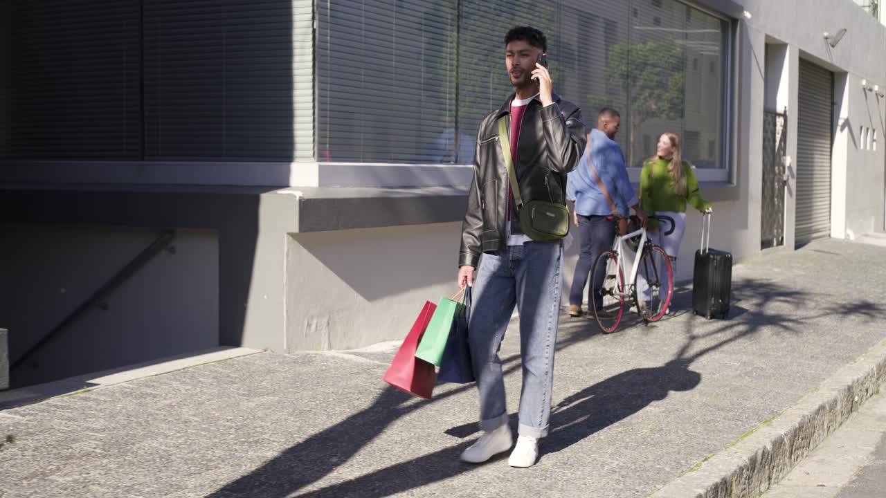 Uomo afroamericano che esce dal negozio, cammina sul marciapiede, parla al telefono e porta tre borse della spesa.