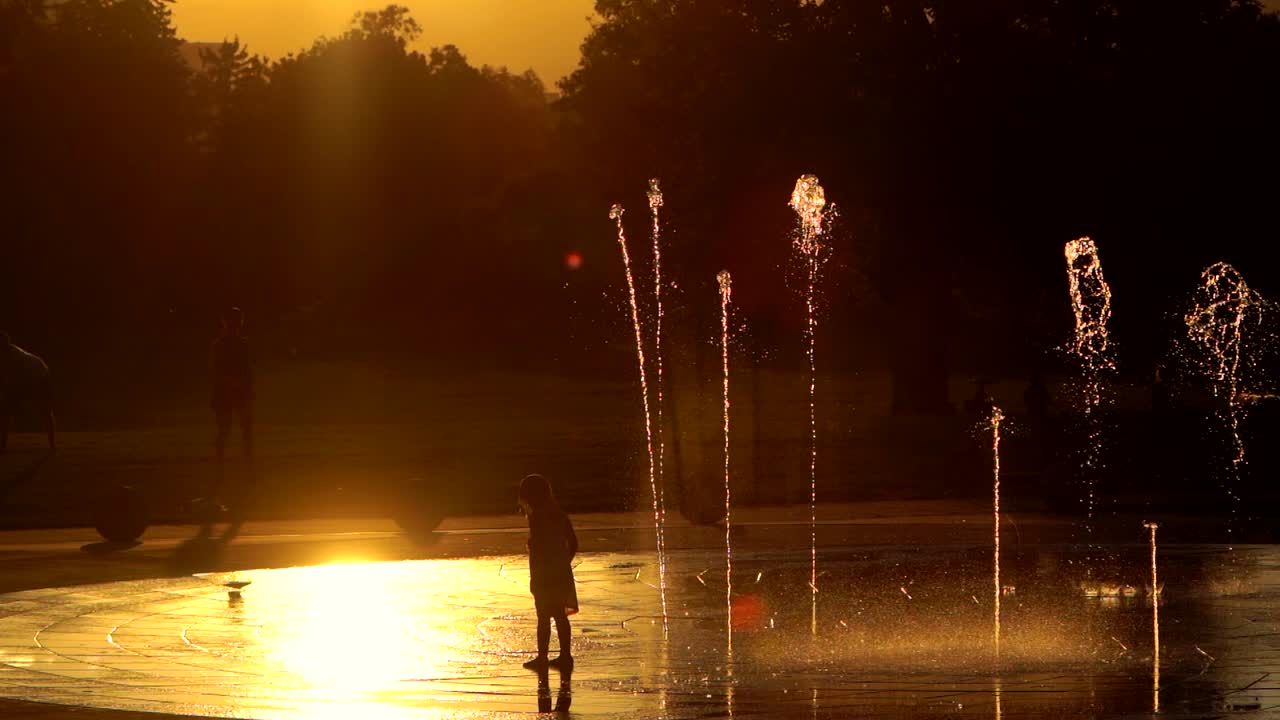 느린 동작으로 일몰을 배경으로 분수대를 가지고 노는 작은 아이