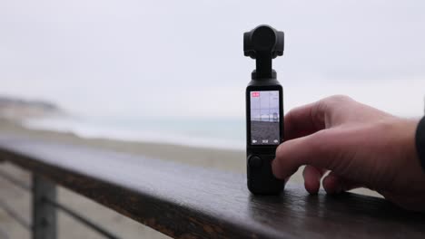 Portable-3-Axis-Stabilized-4K-Handheld-Camera-Being-Turned-On-As-It-Rests-On-Bannister-Beside-Beach-With-Bokeh-Background