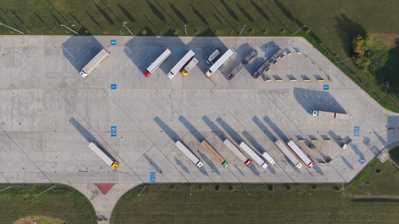 A top-down aerial view of a large truck parking lot showing multiple semi-trailers parked in designated spaces, while one truck moving