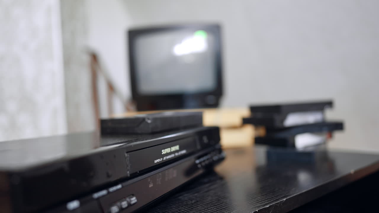 Unrecognized Caucasian man's hand puts the tape into the DVD player. Man presses the buttons on device and screen at backdrop switches on