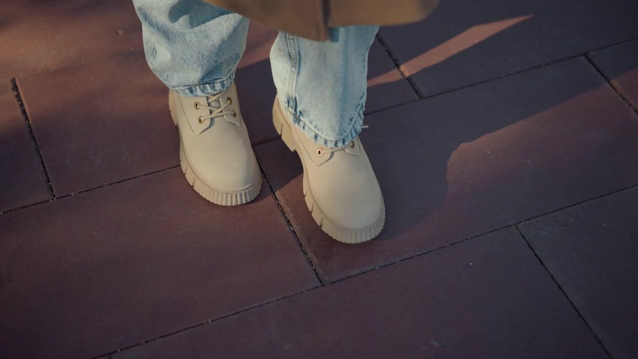 Primo piano dei piedi di una persona con jeans azzurri e scarpe stringate beige che cammina su un marciapiede di mattoni rossi. La scena mostra un movimento costante con illuminazione soffusa che si concentra solo sulla parte inferiore delle gambe.