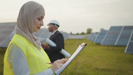 side view of muslim woman in hijab writing on clipboard while standing among rows of solar panels. two multicultural men in helmets standing behind with project plan in hands