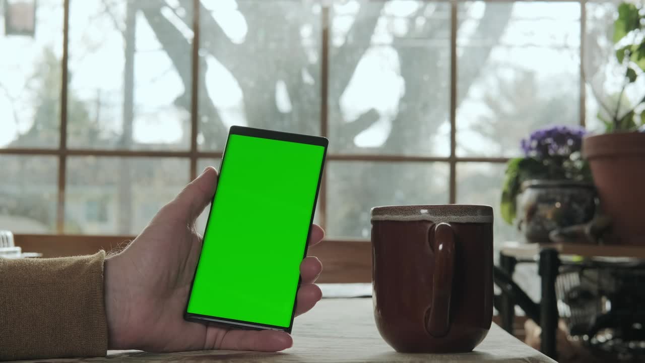 una mano masculina sostiene un teléfono con una pantalla verde de maqueta, y bebe café