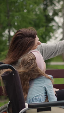 woman enjoys making selfies with little girl in wheelchair after walk in spring park. happy mom and child with disability look in camera of smartphone