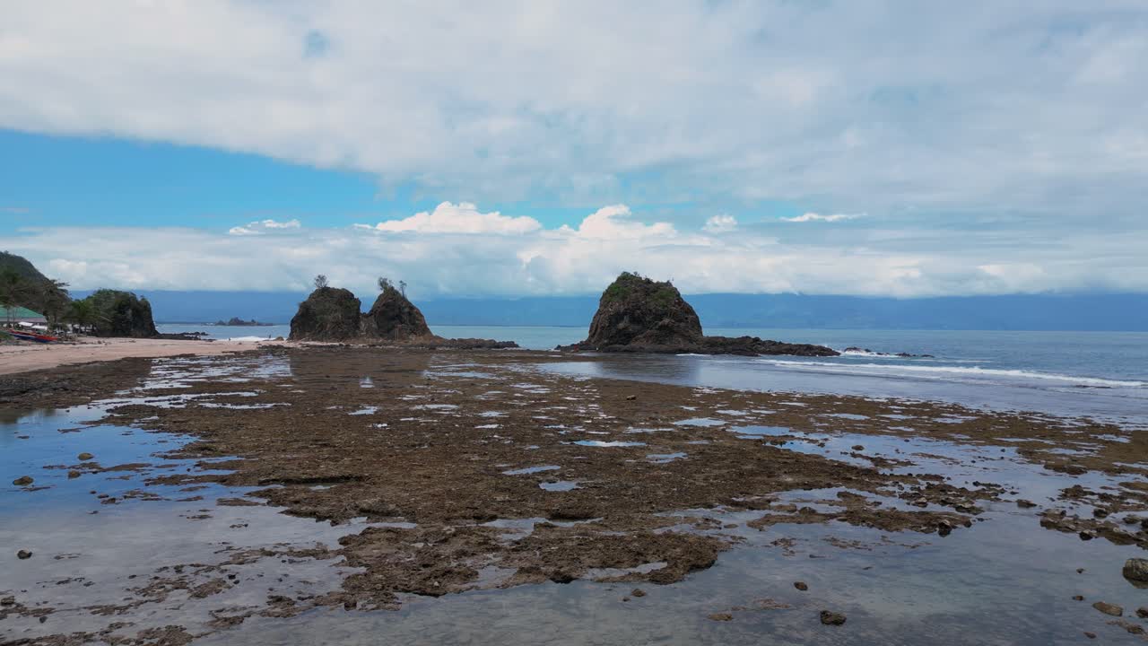 A stunning drone shot captures three small rocky mountains standing proudly near the shore, with the ocean stretching out beyond them.