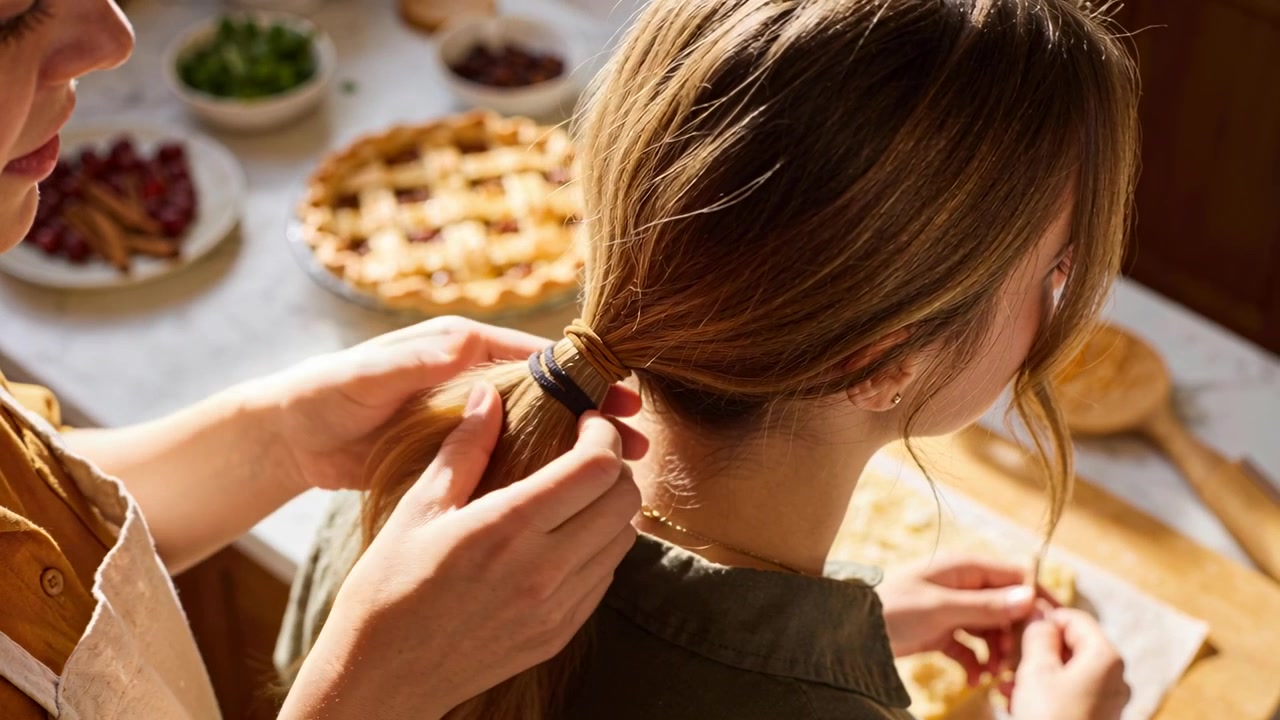 Mamma in maglietta color oliva che si allunga per legare i capelli dell'adolescente in cucina, fissando l'elastico, mentre la figlia modella una torta.
