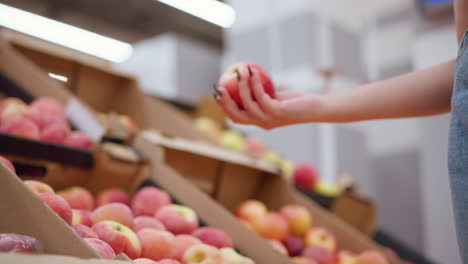 close-up of woman picking red apple from a pile of apples in a box, then dropping it back into the display, focus on hand with black nails interacting with fresh produce at grocery store