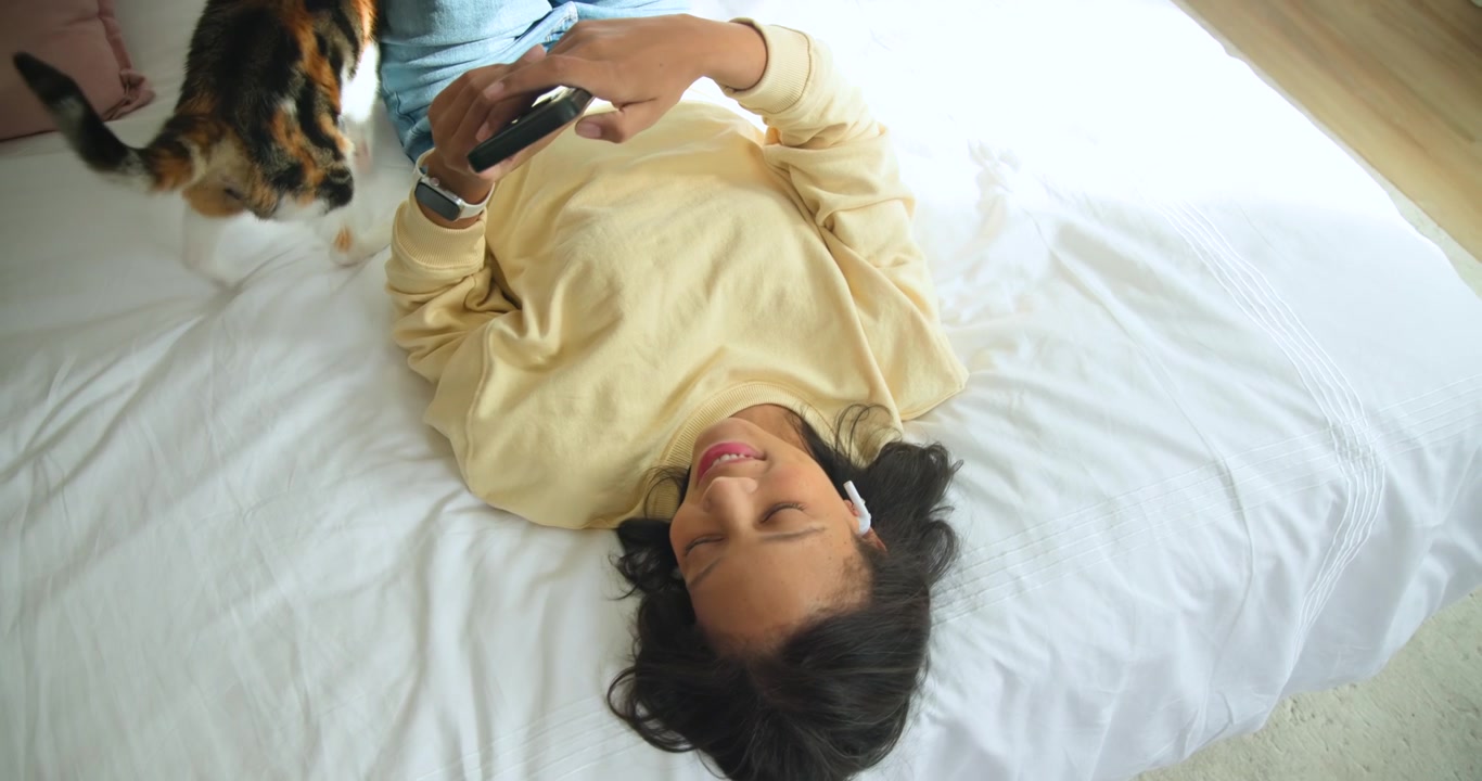 African American woman lying in bed responding to calico cat approaching while holding black phone