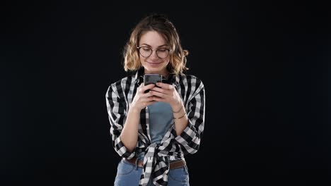 Retrato-De-Una-Mujer-Europea-Concentrada-Vestida-Con-Ropa-Informal:-Camisa-A-Cuadros-Y-Jeans,-Sosteniendo-Un-Teléfono-Inteligente-Y-Escribiendo-Mensajes-De-Texto-O-Charlando,-Aislada-Sobre-Fondo-Negro.-Concepto-De-Emociones,-Amistad,-Tecnología.