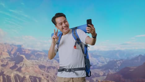 asian male hiker with mountaineering backpack showing peace and thumbs up gesture while taking a selfie while traveling at the top of mountain