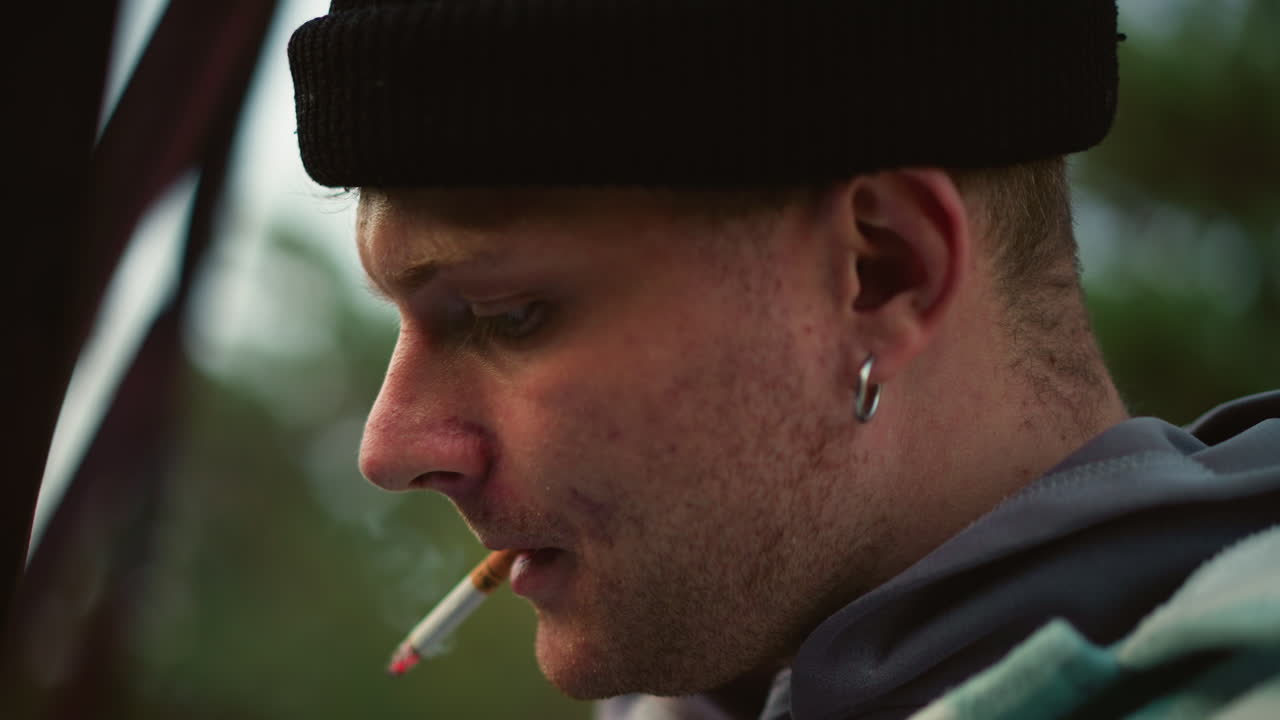 uomo caucasico di profilo che fuma una sigaretta all'aperto, berretto e orecchino, felpa con cappuccio e barba incolta, sguardo pensieroso, crepuscolo al campeggio, grana cinematografica sfocata, stato d'animo riflessivo e tranquillo