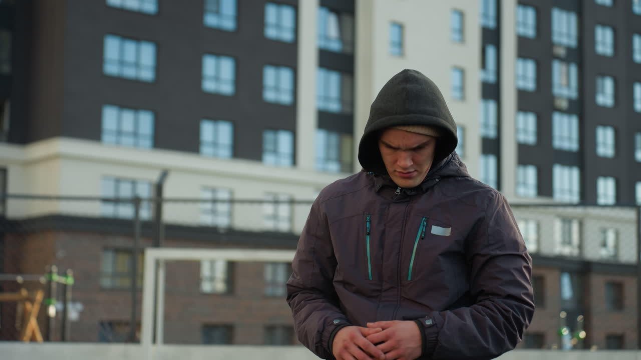 atleta preparándose para una actividad deportiva, girando la cabeza y estirando los brazos mientras lleva una sudadera con capucha en una arena deportiva urbana rodeada de edificios residenciales