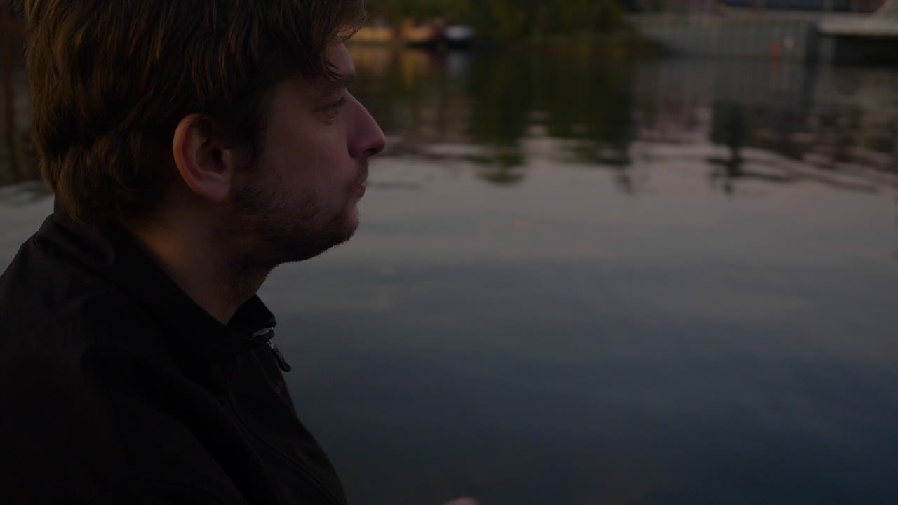 반사되는 호수의 부두에 앉아 감자 칩을 먹는 젊은 남자의 슬로우 모션 측면 프로필