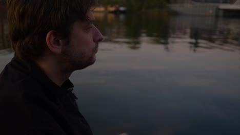 Slow motion side profile close up of a young man sitting on a pier of a reflective lake and eating potato chips
