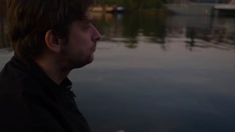 slow motion side profile close up of a young man sitting on a pier of a reflective lake and eating potato chips