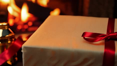 close-up of a gift wrapped in white paper with a red ribbon sitting in front of a fireplace