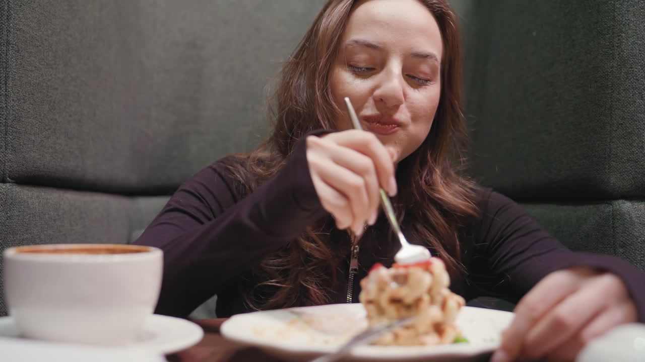 Lady seated in green cafeteria booth smiles as she enjoys waffles with fork, savoring sweet treat beside cup of latte on wooden table, capturing cozy and cheerful breakfast moment in modern setting