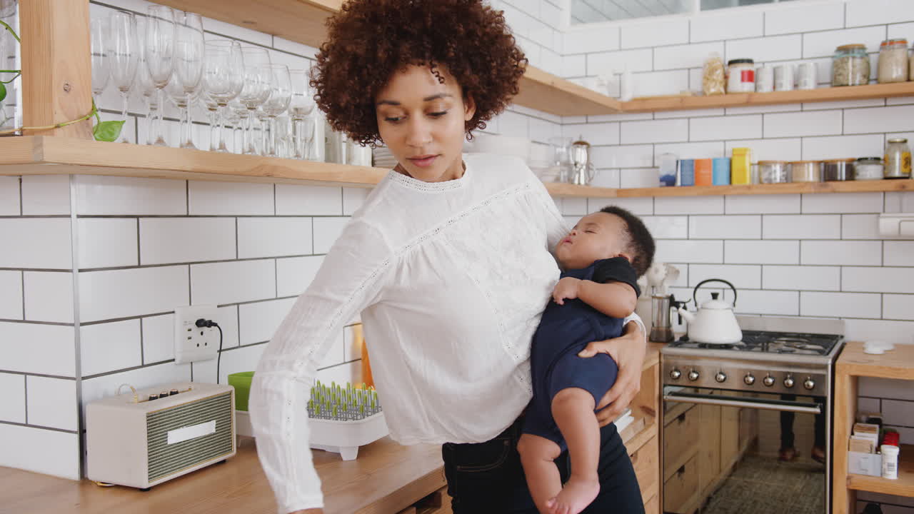 Mère multitâche tenant son bébé endormi tout en nettoyant et en travaillant sur un ordinateur portable dans la cuisine