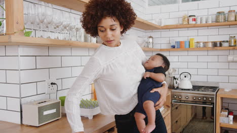 multi-tasking mother holds sleeping baby son whilst cleaning and working on laptop in kitchen