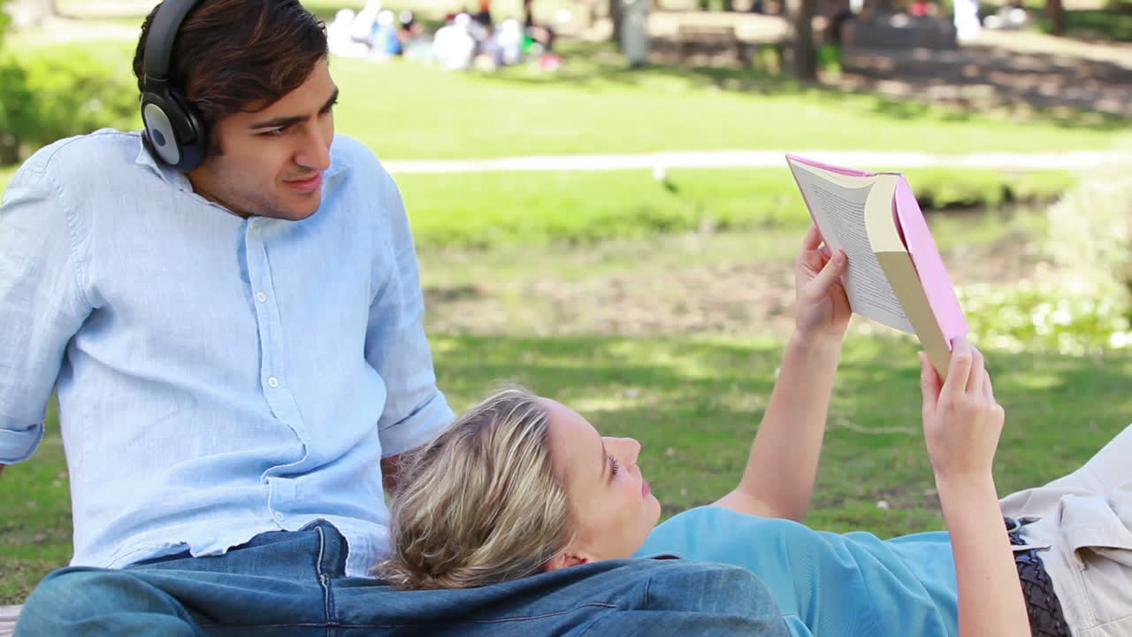 한 남자가 헤드폰을 들으면서 한 소녀가 그의 다리에 누워서 책을 읽습니다.