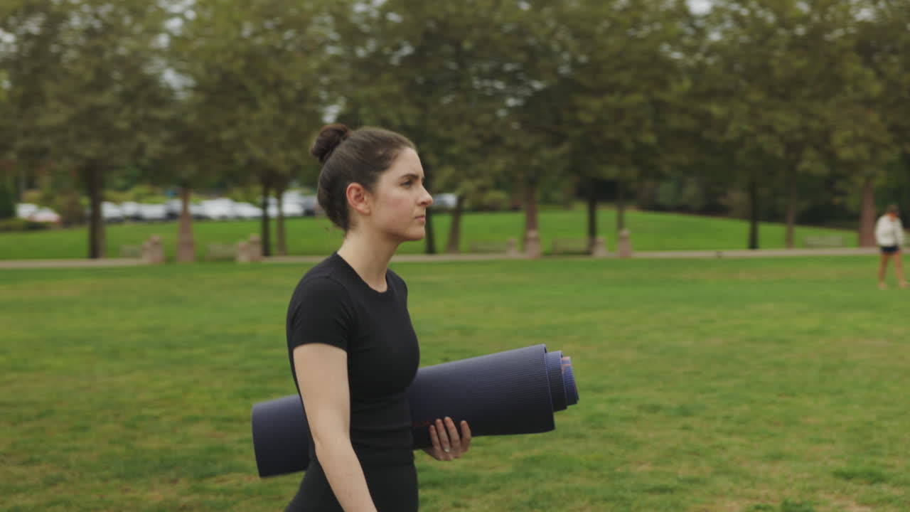 joven mujer blanca en forma atlética con atuendo negro de yoga que lleva una colchoneta de yoga a través de un parque público en seattle, washington
