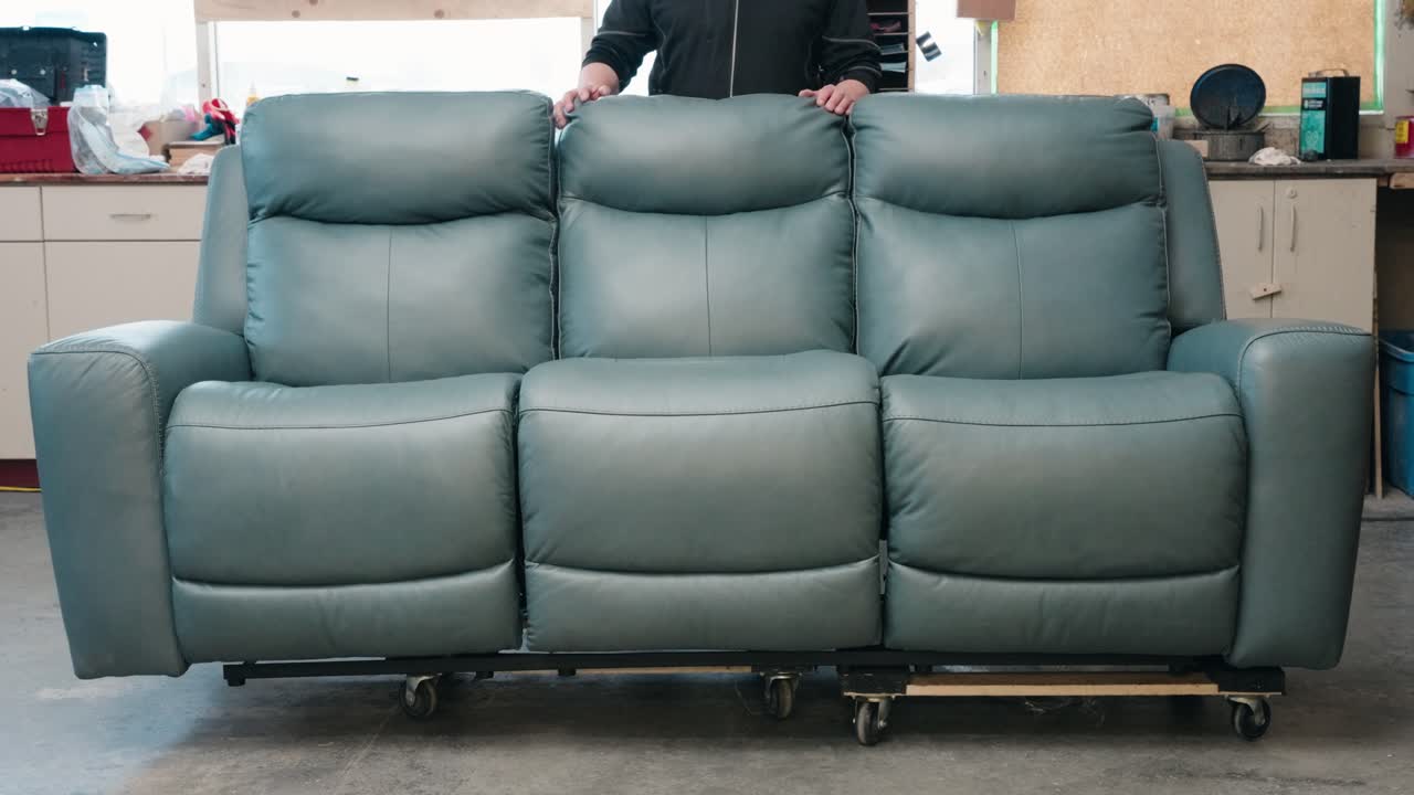 Worker in carpentry shop stands behind blue sofa on rollers, ready to work