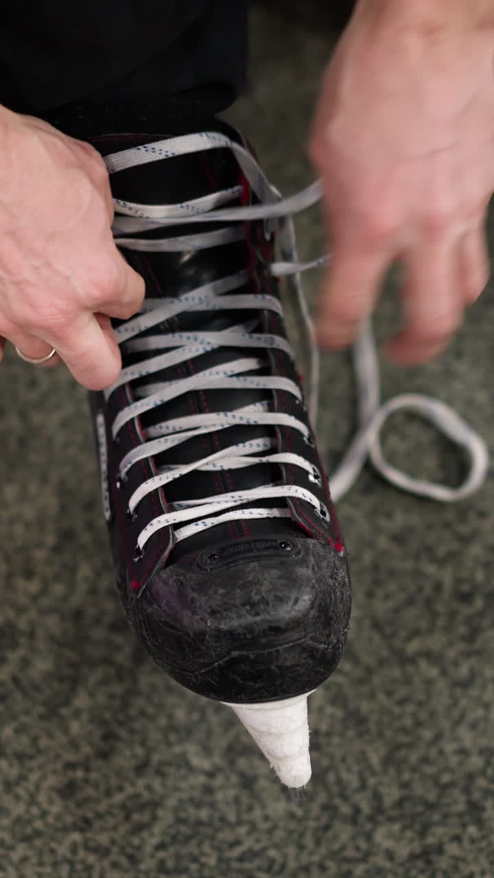 Close-up of hands loosening the lace of a black ice skate with red accents on a gray carpeted floor