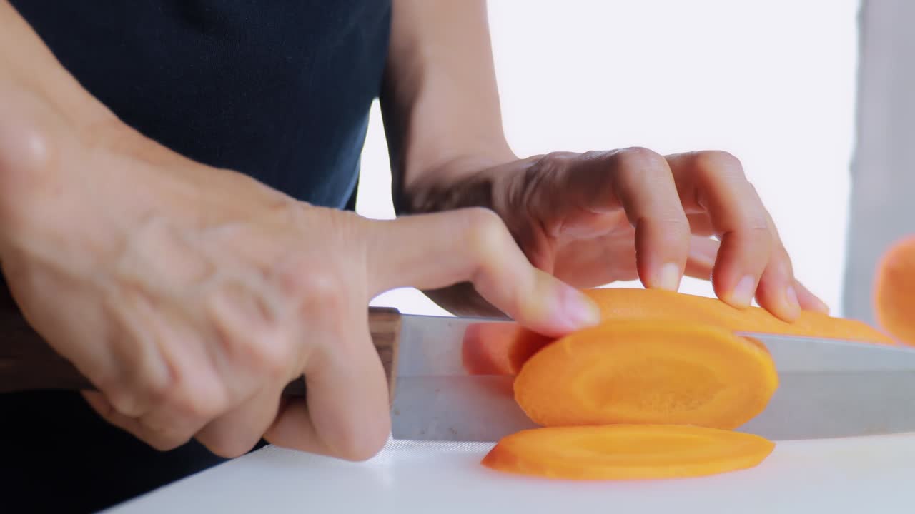 4K Kadın Eli Kesme Tahtasında Taze Havuç Kesiyor. Kadın eli, kesme tahtasında çiğ havuçları bıçakla daireler halinde dilimliyor. Sağlıklı beslenme konsepti.