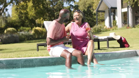 senior friends sitting by pool, laughing and enjoying drinks on sunny day