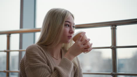 smiling lady sipping tea, looking outside with a relaxed expression, with blurred cityscape in background, seated comfortably in modern cafe, enjoying peaceful moment with soft lighting