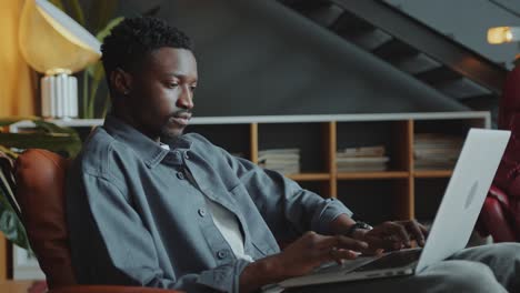 Focused Black Man Sitting in Armchair and Surfing the Net on Laptop in Office