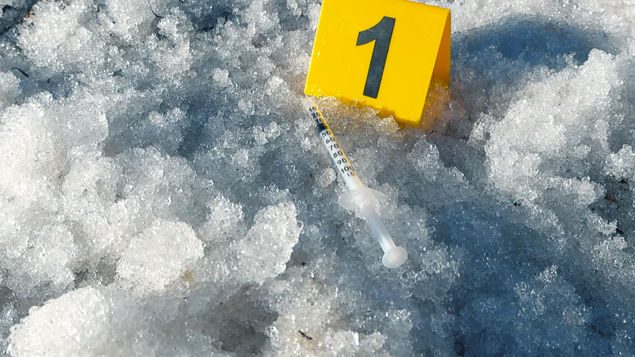 Investigación de la escena del crimen con un marcador amarillo junto a una jeringa usada, hallada como evidencia sobre la nieve húmeda y derritiéndose, sugiriendo consumo de drogas, sobredosis o una investigación de actividad ilegal.