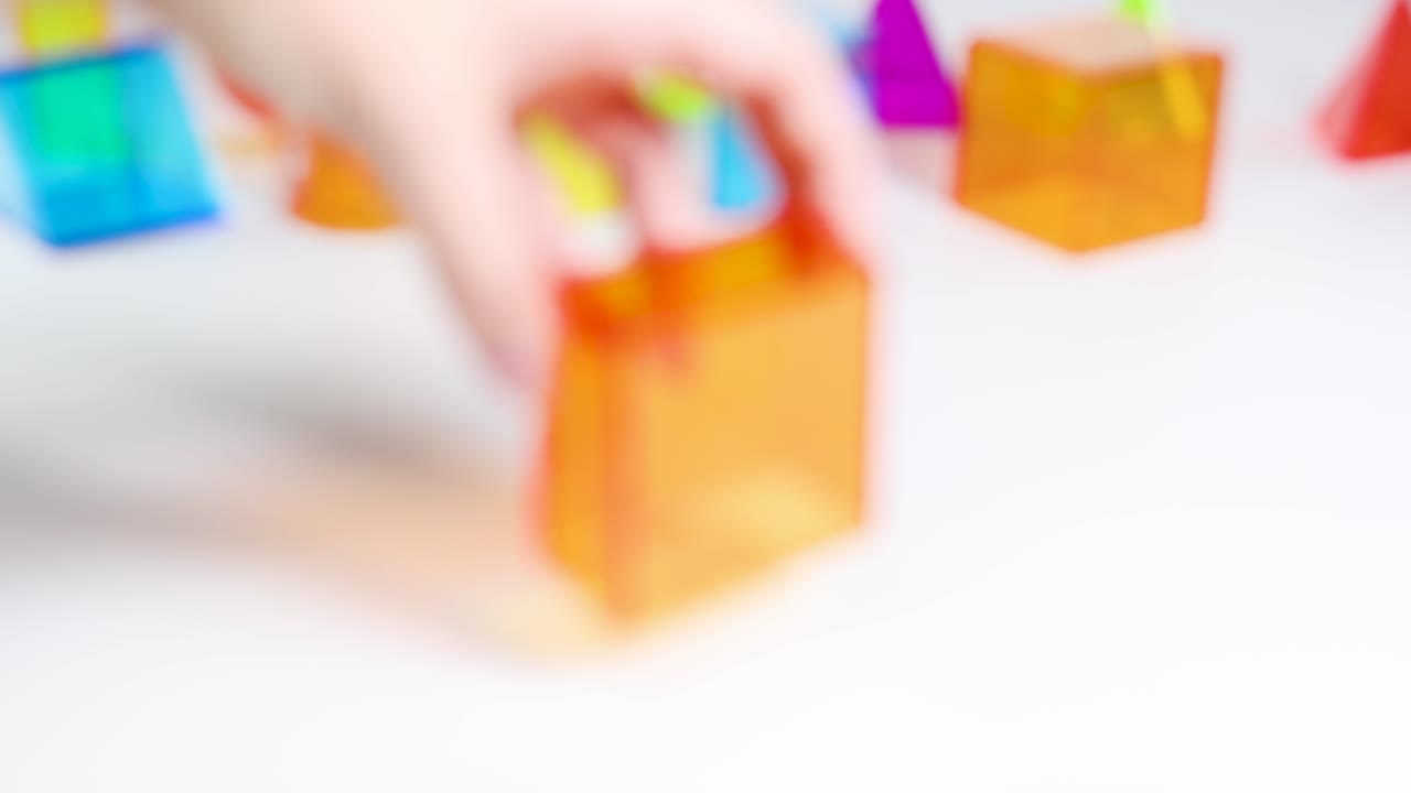 Hands manipulate vibrant 3D geometric shapes on a white surface, exploring form and color in a bright, educational setting