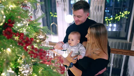 lovely married couple with an infant baby. family of three stand near the big christmas tree looking at decorations.