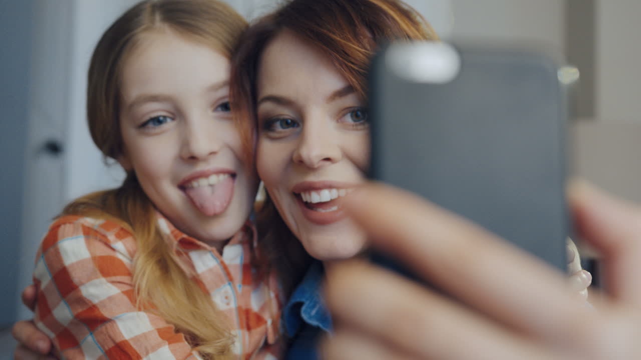 Portrait shot of the beautiful woman with young daughter making selfies on the smart phone, showing their tongues, hugging and laughing. Close up. Inside